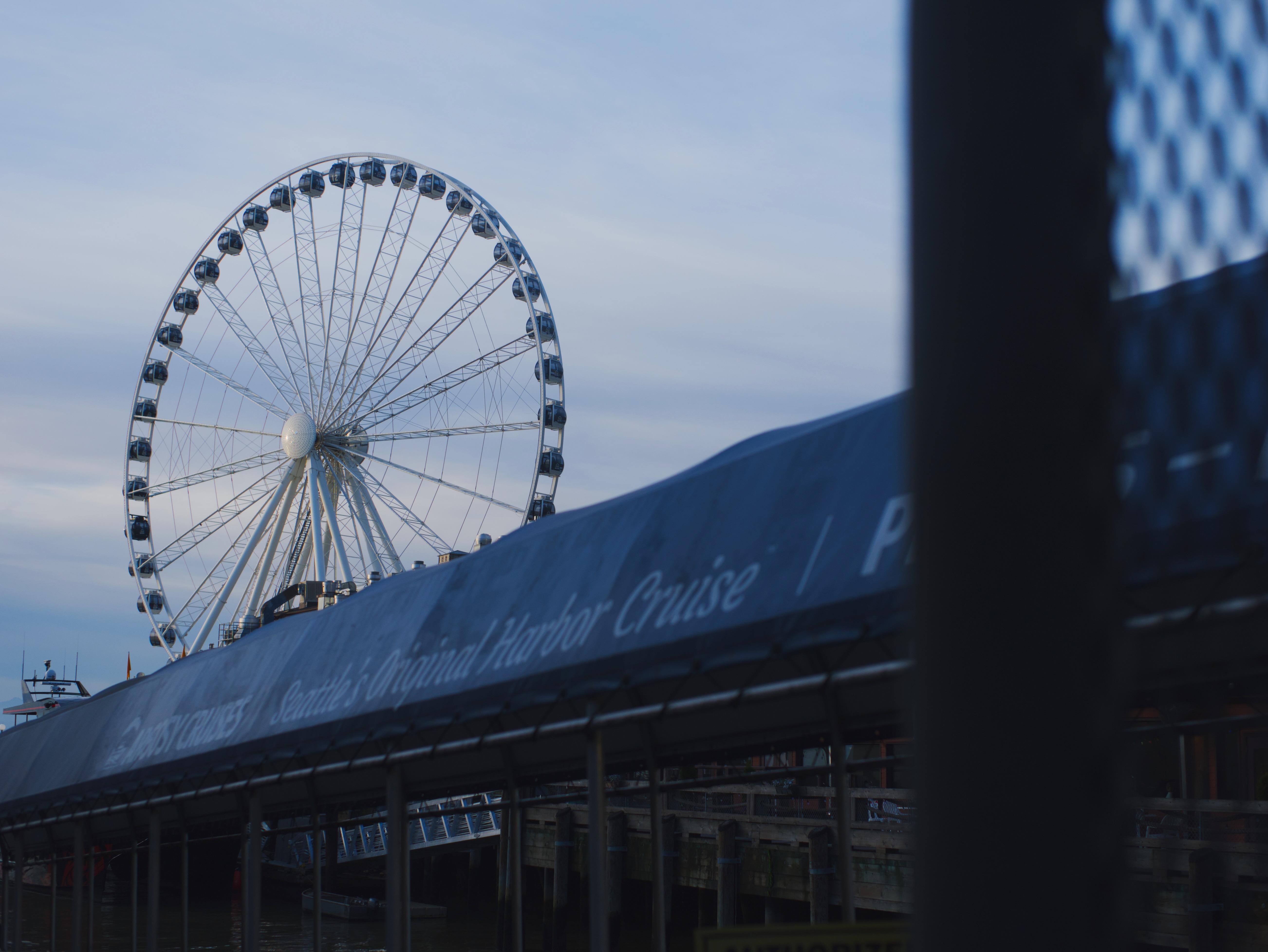 A large Ferris wheel rises behind a waterfront building with 'Seattle Harbor Cruise' signage at dusk.