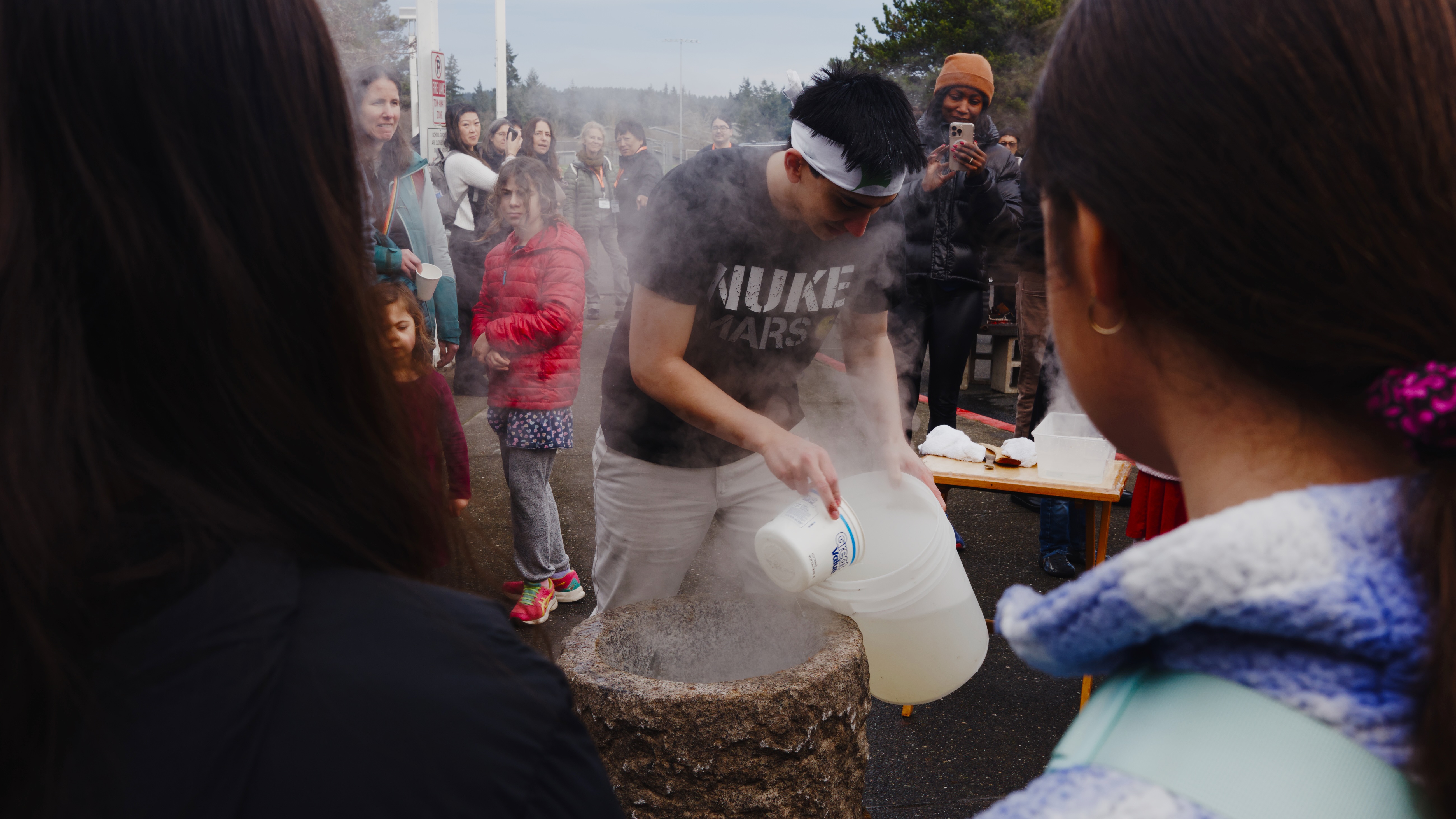 A man wearing a headband pours water into a traditional stone mortar (usu) during a mochi-pounding demonstration as a crowd watches outdoors.