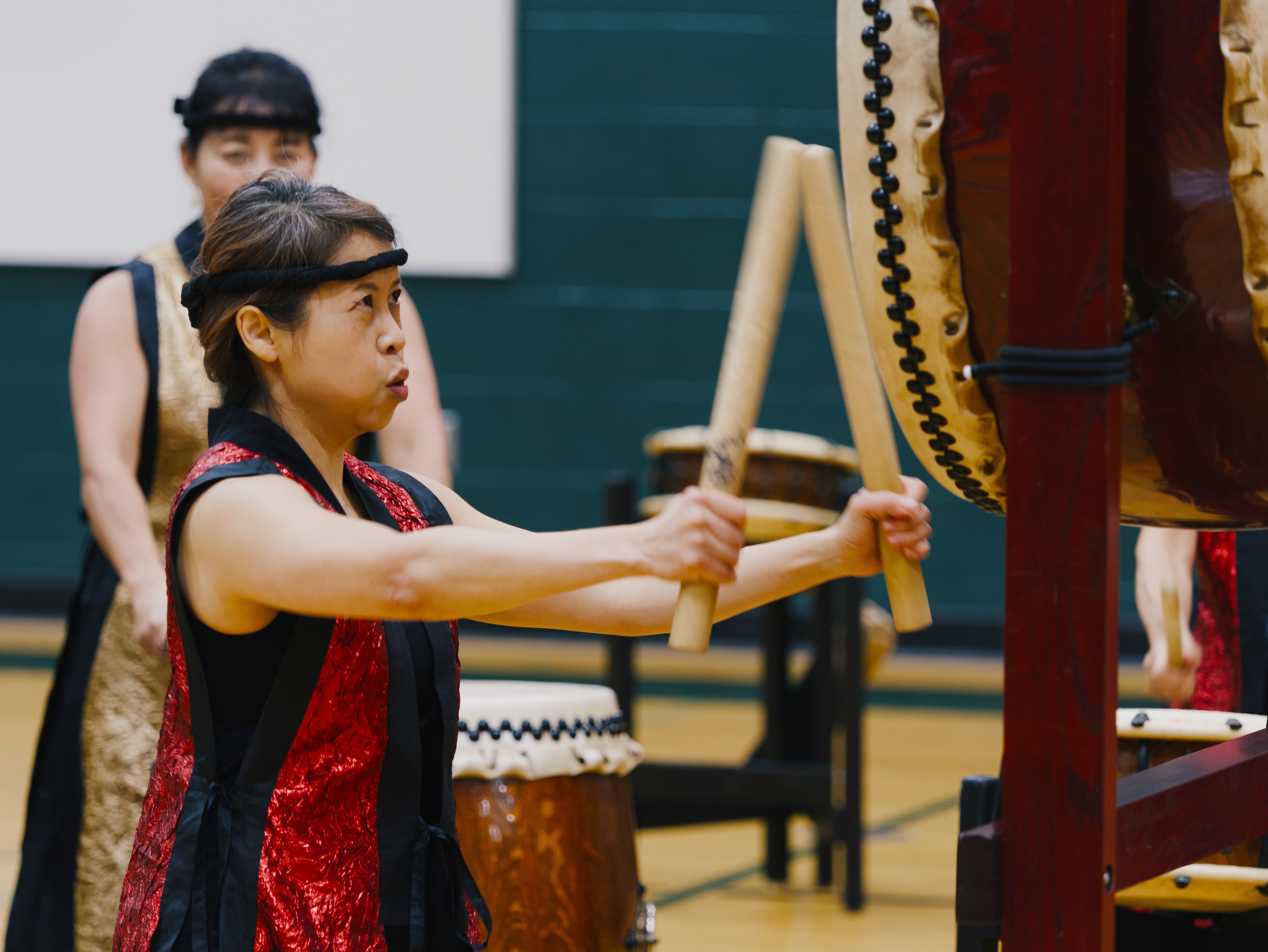 A taiko drummer in a red and black vest performs with large wooden bachi sticks on a traditional Japanese drum during a group performance.